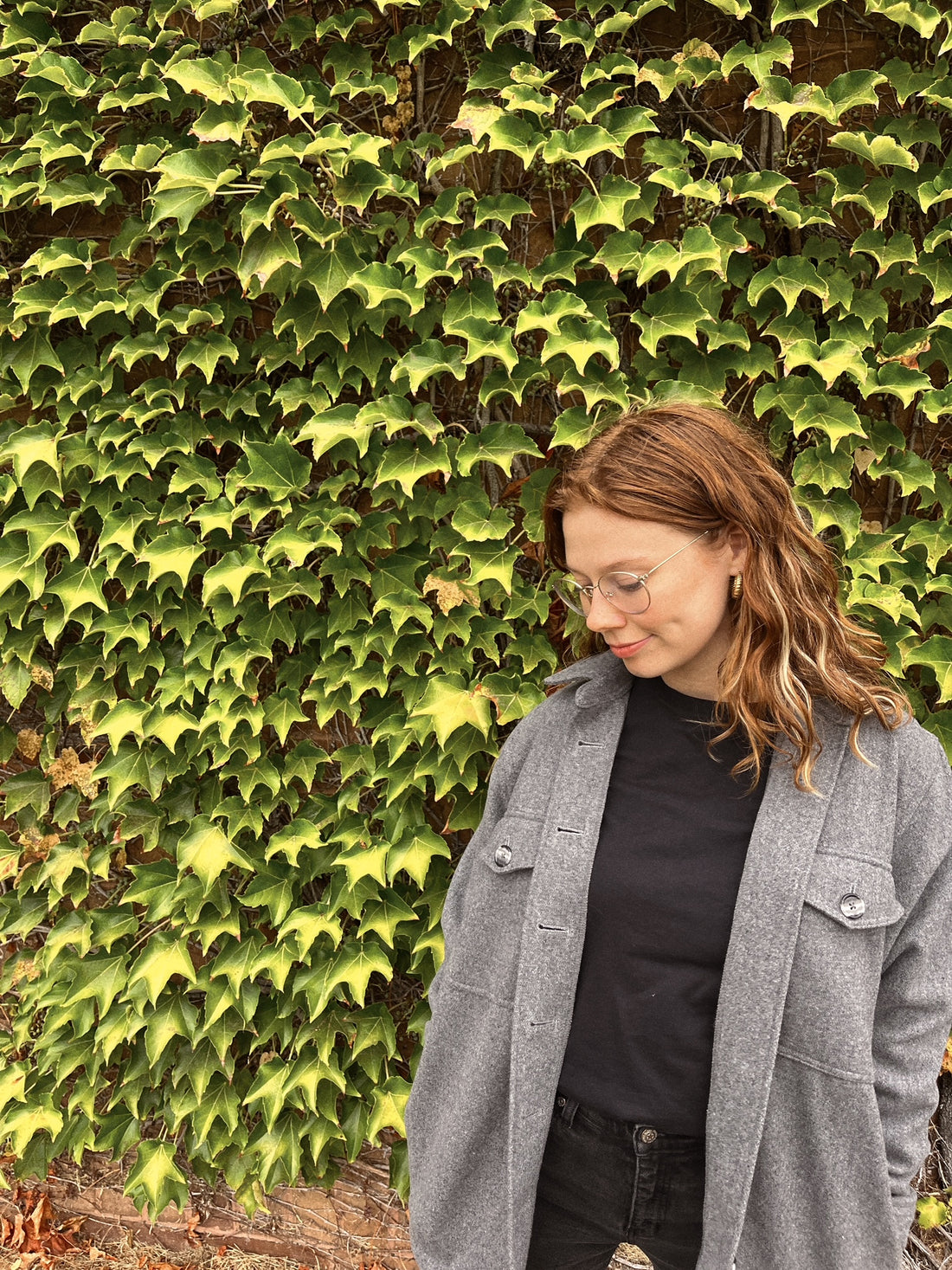 The Shay Shacket/Jacket in "Grey" is featured in this image with Millie standing in front of a green wall of leaves.
